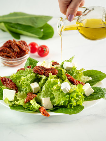 Salad of sun-dried tomatoes, feta cheese and salad leaves on white table, vertical. Olive oil pouring on salad. High key light. Close up. Selective focus. Copy space. Side viewの写真素材