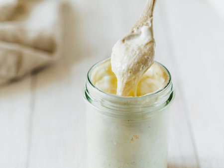Ready for making bread wheat sourdough starter dripping from wooden spoon. Glass jar with sourdough starter 100 percent hydration on white wooden background. Copy space. Homemade breadmaking conceptの写真素材