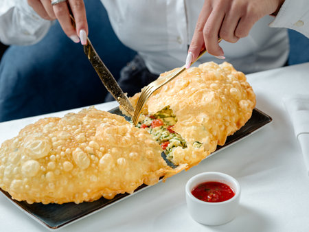 Fried cheburek with feta cheese, on the restaurant table. Female hands cutting big cheburek with...