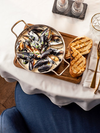 Delicious black mussels in creamy cheese and garlic sauce. Mediterranean black mussels in metal pan on wooden cutting board with grilled bread on restaurant table background, top viewの写真素材