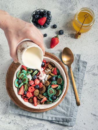 Trendy food - pancake cereal. Milk pouring on heap of multicolor mini cereal pancakes in bowl. Tiny cereal pancakes with berries in craft plate over gray cement background. Vertical. Top view,flat layの写真素材