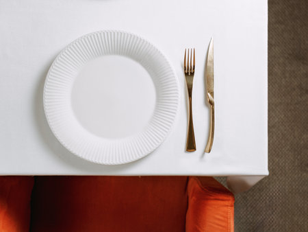 Top view of table in restaurant or dinning room with white round empty plate and gold cutlery. White tablecloth, red chair and table setting as background for your text or design. Copy spaceの写真素材