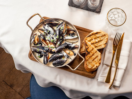 Delicious black mussels in creamy cheese and garlic sauce. Mediterranean black mussels in metal pan on wooden cutting board with grilled bread on restaurant table background, top viewの写真素材