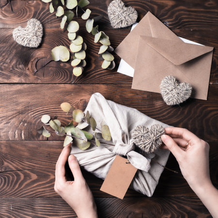 Zero waste, Valentines Day eco-friendly gift wrapping in Furoshiki style with dry eucalypt. Hands with gift box in cloth wrapping with empty craft label on brown wooden background. Top view, flat layの写真素材