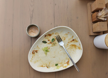Dirty plate after dinner. Top view of dirty white plate with fork over wooden tabletop. Dirty plate after eating salad with sauce on the dining table. Copy spaceの写真素材