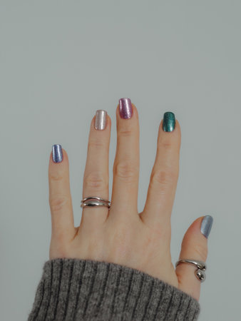 Colorful shiny manicure with holographic sparkle and metallic pigment. Female hand in gray sweater with multicolored nails on white wall background. Copy space. Verticalの写真素材