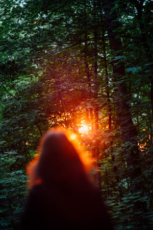 Backlit Silhouette Of Woman In Forest With Sunbeams Through Tree Branch In Sunset Light. Peaceful nature scene of woman standing in sunset light with sun rays through green leaves, back viewの写真素材