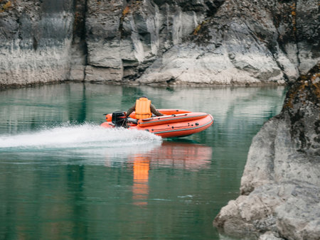 Orange inflatable motorboat with outboard engine navigates azure mountain river. Man in bright life jacket steers boat through azure water amid rocky cliffs. Visible water marks on rocks in backgroundの写真素材