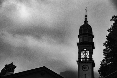 lonely bell tower stands out against the blue sky of a summer evening with the bells illuminated by a warm light and the shadow of the roof of a house.の写真素材