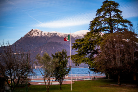 Italian flag flutters on a flagpole in a lakeside park with trees and green lawnsの写真素材