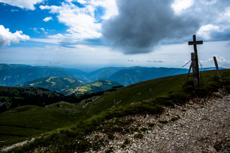 Dramatic mountain view from the Asiago Plateau, Italy, with a wooden cross on a rocky path overlooking green valleys, forests, and distant blue peaks under a moody sky.の写真素材