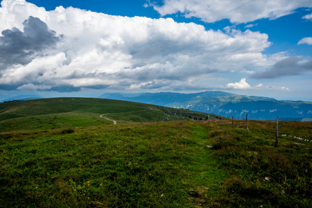Vast alpine meadow with winding trail and wooden fence under dramatic clouds in Asiago, Italy....