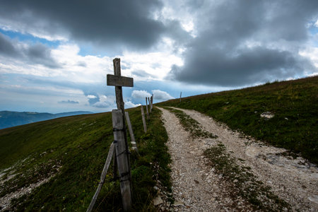 Gravel mountain path lined with wooden posts under dramatic clouds in Asiago, Italy. Rustic alpine trail with moody sky and wild sceneryの写真素材