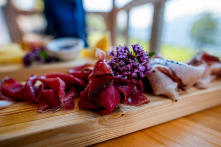 lose-up of Italian cured meats and cheese on wooden board with purple wildflowers. Traditional alpine food platter with natural light and blurred background. Rustic gourmet dining concept.の写真素材