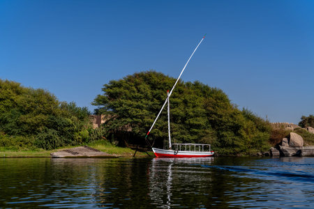 Single felucca sailboat moored along the Nile River in Aswan, Egypt, framed by lush greenery and...
