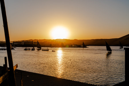 Golden sunset over the Nile River in Aswan, Egypt, with traditional sailboats crossing calm water....