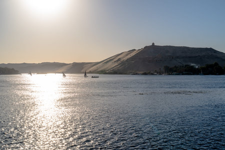 Sailboats gliding across the Nile in Aswan, Egypt, with strong sunlight reflections and desert hills...