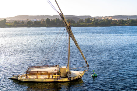Traditional felucca boat anchored on the Nile in Aswan Egypt with calm water and green riverbank in...