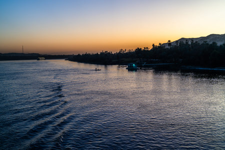 Twilight view of the Nile River in Aswan with smooth water, palm-lined shores, distant hills, and...