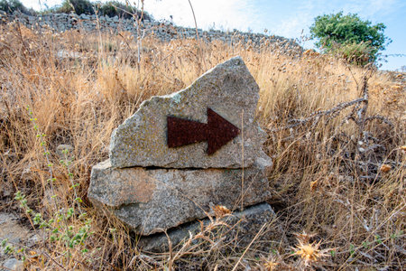 Stone trail marker with a metal arrow embedded in rock, set in dry grass and natural terrain in...