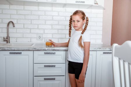 Little girl drinks fresh orange juice from a glass cup in the kitchenの写真素材