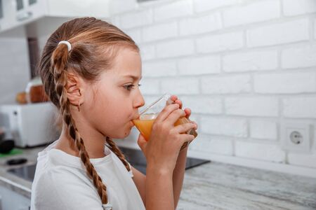 Little girl drinks fresh orange juice from a glass cup in the kitchenの写真素材