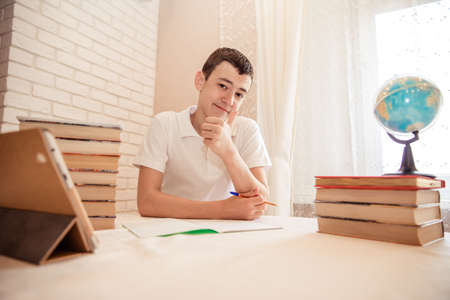 student sits at the table at the bookside with a pen in his hands and looks at the camera. A tired...