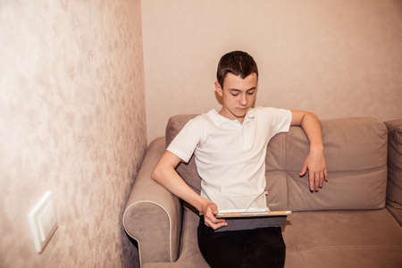 Teenager boy in white T-shirt sits on sofa with laptop. Home education, self education by kids.