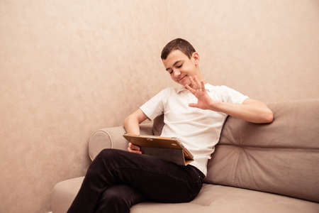 Teenager boy in white T-shirt sits on sofa with laptop. Home education, self education by kids.