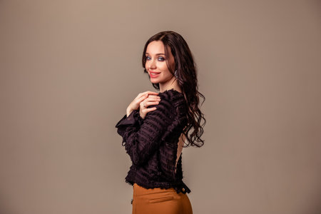 beautiful slender young girl with black long hair on a one-ton beige background. woman in brown trousers and black blouse with open back. Copy spaceの写真素材