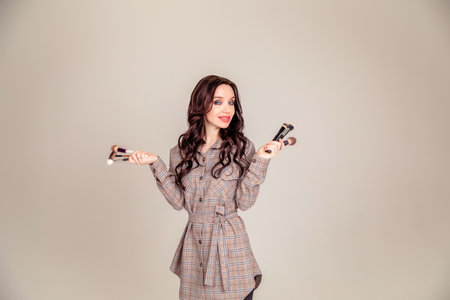 Young beautiful woman holding makeup brushes in her hands and posing on gray background at studio. Visagiste, makeup artist. Copy spaceの写真素材