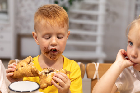 little red boy eats a croissant with chocolate. boy dirty his mouth and face in chocolateの写真素材