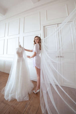 bride morning. a girl in a white robe and veil stands next to a white wedding dressの写真素材