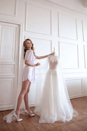 bride morning. a girl in a white robe and veil stands next to a white wedding dressの写真素材