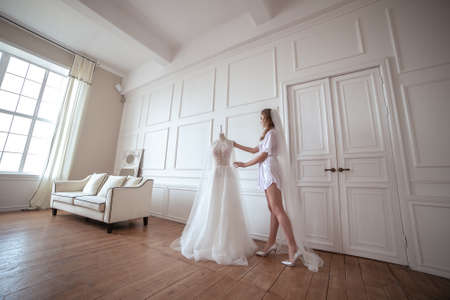 bride morning. a girl in a white robe and veil stands next to a white wedding dressの写真素材