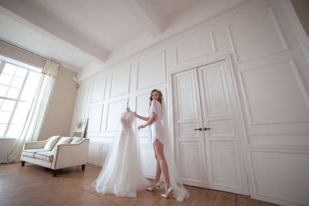 bride morning. a girl in a white robe and veil stands next to a white wedding dressの写真素材