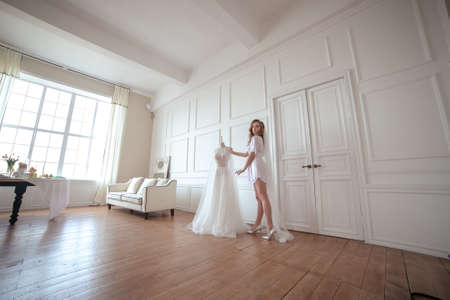 bride morning. a girl in a white robe and veil stands next to a white wedding dressの写真素材