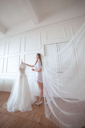 bride morning. a girl in a white robe and veil stands next to a wedding dressの写真素材