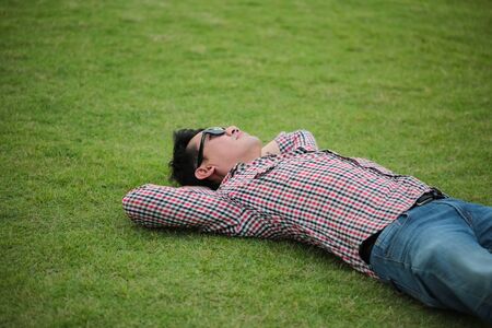 Man lying on green grass wearing check shirt, blue jeans at a park and looking on the sky on summer dayの写真素材
