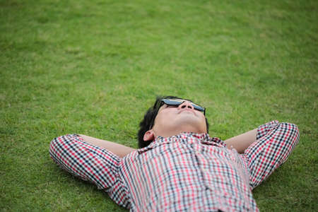 Man lying on green grass wearing check shirt, blue jeans at a park and looking on the sky on summer dayの写真素材
