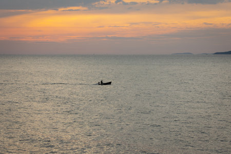 silhouette lone boat cuts through the calm waters as the sun begins its descent, casting a warm glow across the horizon. The serene scene captures the tranquility of the ocean and the beauty of a golden sunset.の写真素材