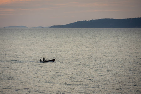 silhouette lone boat cuts through the calm waters as the sun begins its descent, casting a warm glow across the horizon. The serene scene captures the tranquility of the ocean and the beauty of a golden sunset.の写真素材