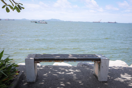 Concrete bench near greenery facing the ocean, with boats scattered across the water under a sunny sky and light clouds.の写真素材