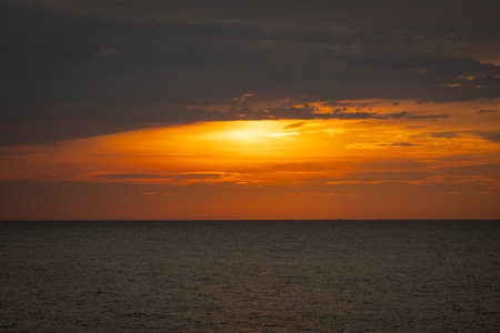 The sun dips below the horizon, casting a golden glow over the calm sea. A gentle breeze ruffles the water as a lone sailboat glides peacefully across the bay. Seagulls cry overhead, adding to the tranquil atmosphere.の写真素材
