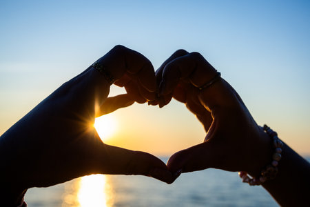 Silhouetted hands forming a heart shape against a golden sunset over the ocean, with sunlight reflecting on the calm water.の写真素材