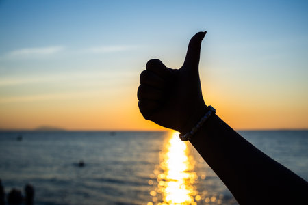 silhouetted hand giving a thumbs-up gesture against a vibrant sunset over the ocean, with golden sunlight reflecting on the water.の写真素材
