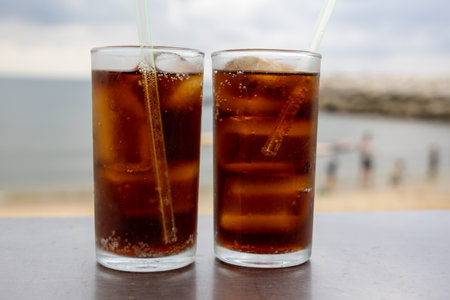 glass of iced cola sits on a table by the sea, with a cloudy sky backdrop. The drink's bubbles and condensation are visible, highlighting its refreshing quality. The beach setting creates a relaxed, summer vibe.の写真素材
