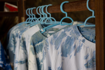 close-up shot of tie-dye t-shirts hanging on light blue plastic hangers. The shirts feature a soft, pastel tie-dye pattern. The hangers are neatly arranged on a wooden rackの写真素材