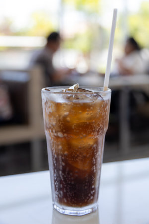 A glass of ice-cold cola sits on a table, condensation glistening on the glass. The bubbles fizz...
