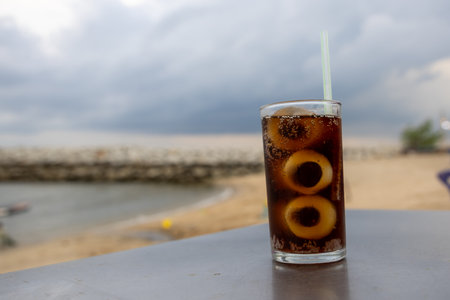 glass of iced cola sits on a table by the sea, with a cloudy sky backdrop. The drink's bubbles and...